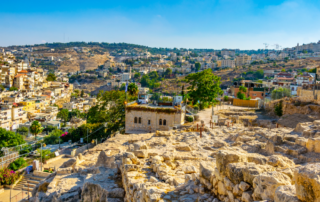 Aerial view of the City of David with a small stone structure in the foreground and a cityscape in the background, with houses and buildings covering the hilly landscape under a clear blue sky.